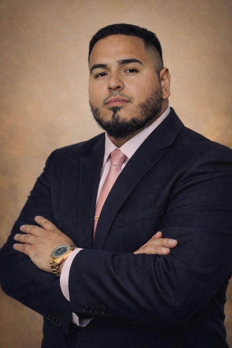 Professional headshot of a man in a dark navy blazer and pink shirt with arms crossed, wearing a gold watch against a beige background.